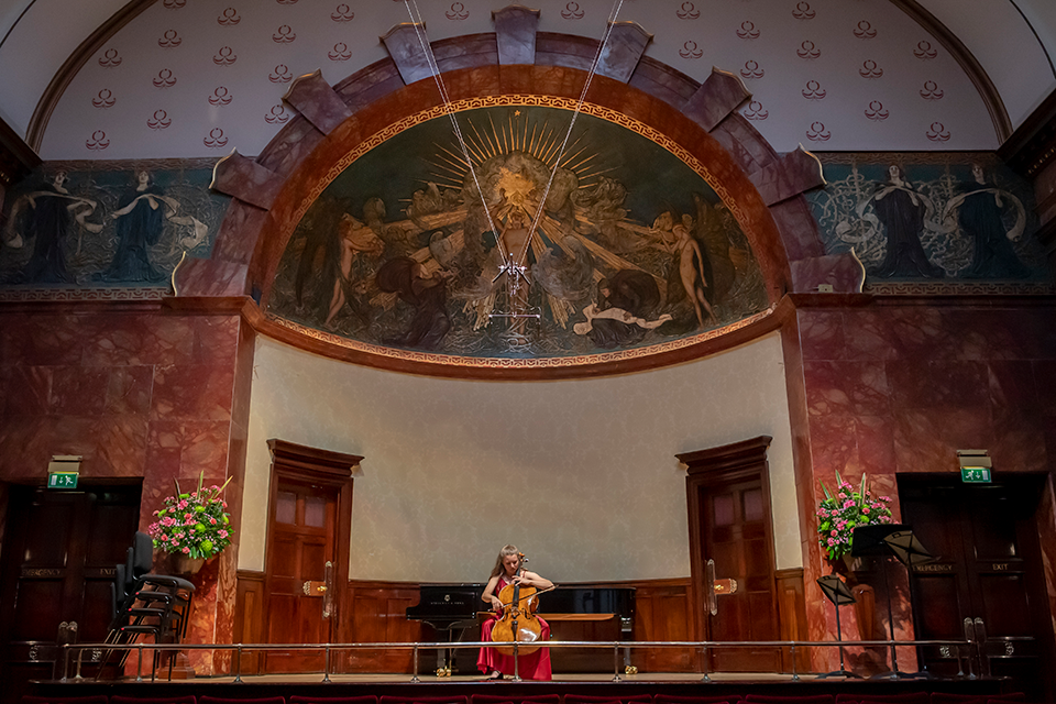 A student cellist performing on the stage of an ornate chamber music venue 